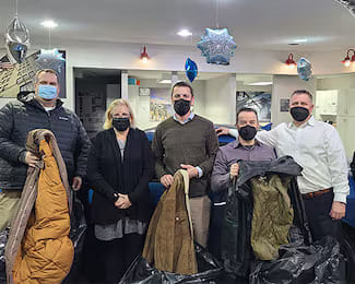 Five individuals holding donated winter coats and large bags filled with donations indoors.