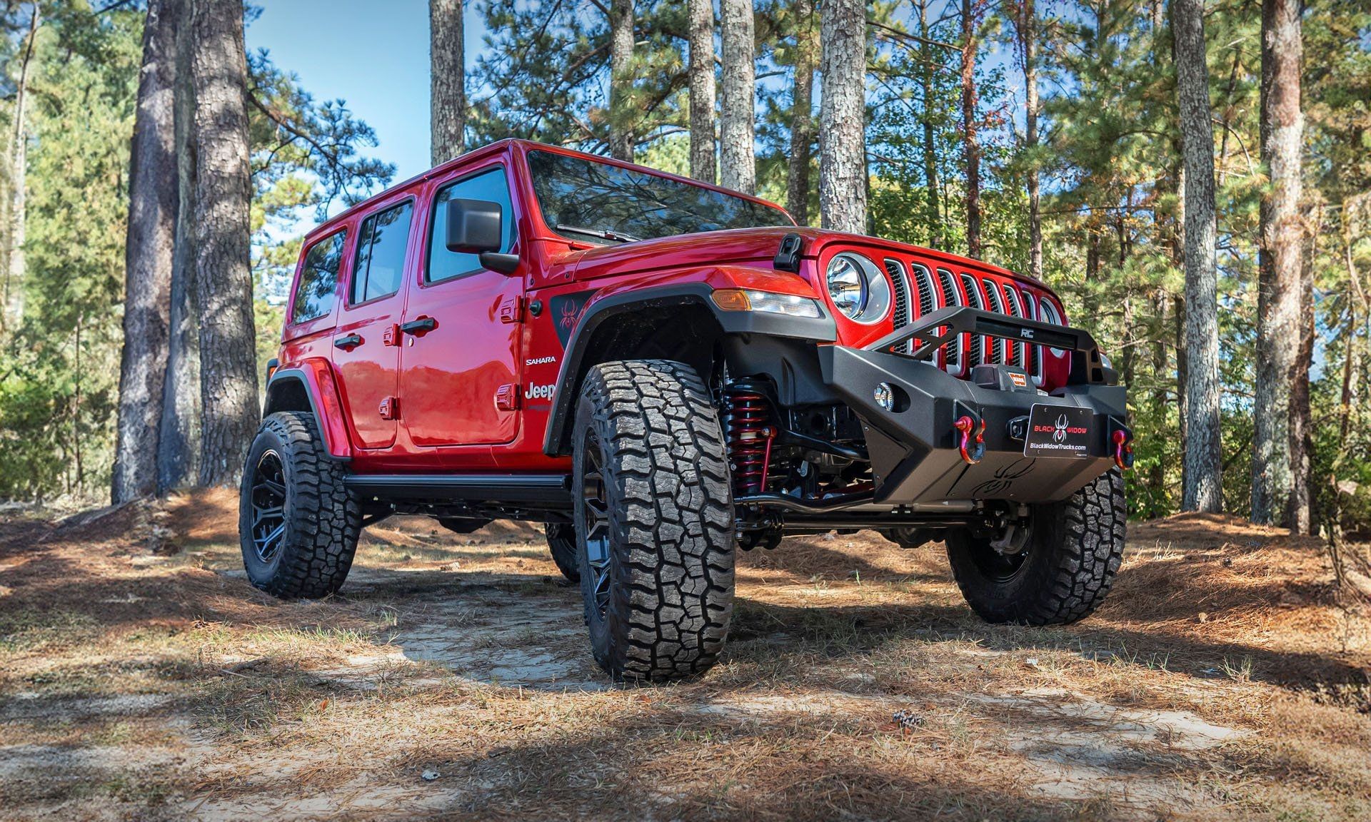 A red Jeep Wrangler Black Widow Edition parked amidst trees in a wooded area.
