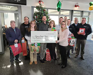 Group of Tri City Cars employees presenting a donation check to Lydia's House, holding gift bags in front of a decorated Christmas tree in a showroom.