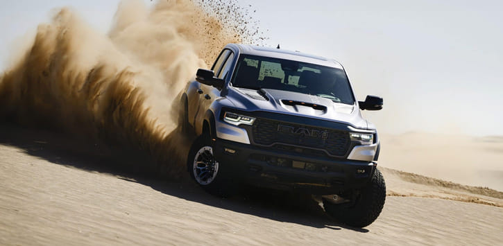 A head-on angle of a silver 2025 Ram 1500 driving off-road on a hill in the desert, with dust and sand billowing from its tires.