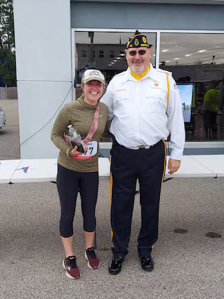 Young woman wearing race bib number 77 poses smiling next to a man in an American Legion uniform outdoors at an event.
