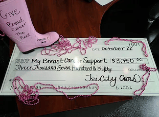 Donation check from Tri City Cars to My Breast Cancer Support, displayed on a desk with pink beads and a pink boot reading 'Give Breast Cancer The Boot.'