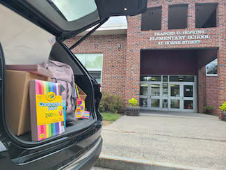 Open trunk of an SUV filled with donated school supplies like construction paper and backpacks, parked in front of the brick Frances G. Hopkins Elementary School building.