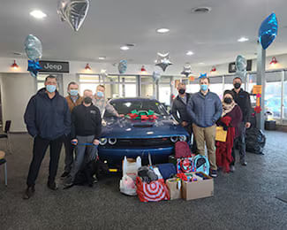 Tri City Cars employees posing in the dealership showroom around a blue Dodge Challenger topped with a bow, with numerous donated gifts and backpacks arranged in front.