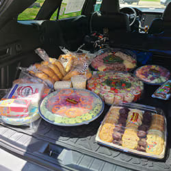 Cargo area of a car filled with numerous large catering platters of food, including sandwiches, cookies, fruit, vegetable, and deli trays.