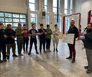 Man presenting a check to a group of applauding students in automotive garage setting.