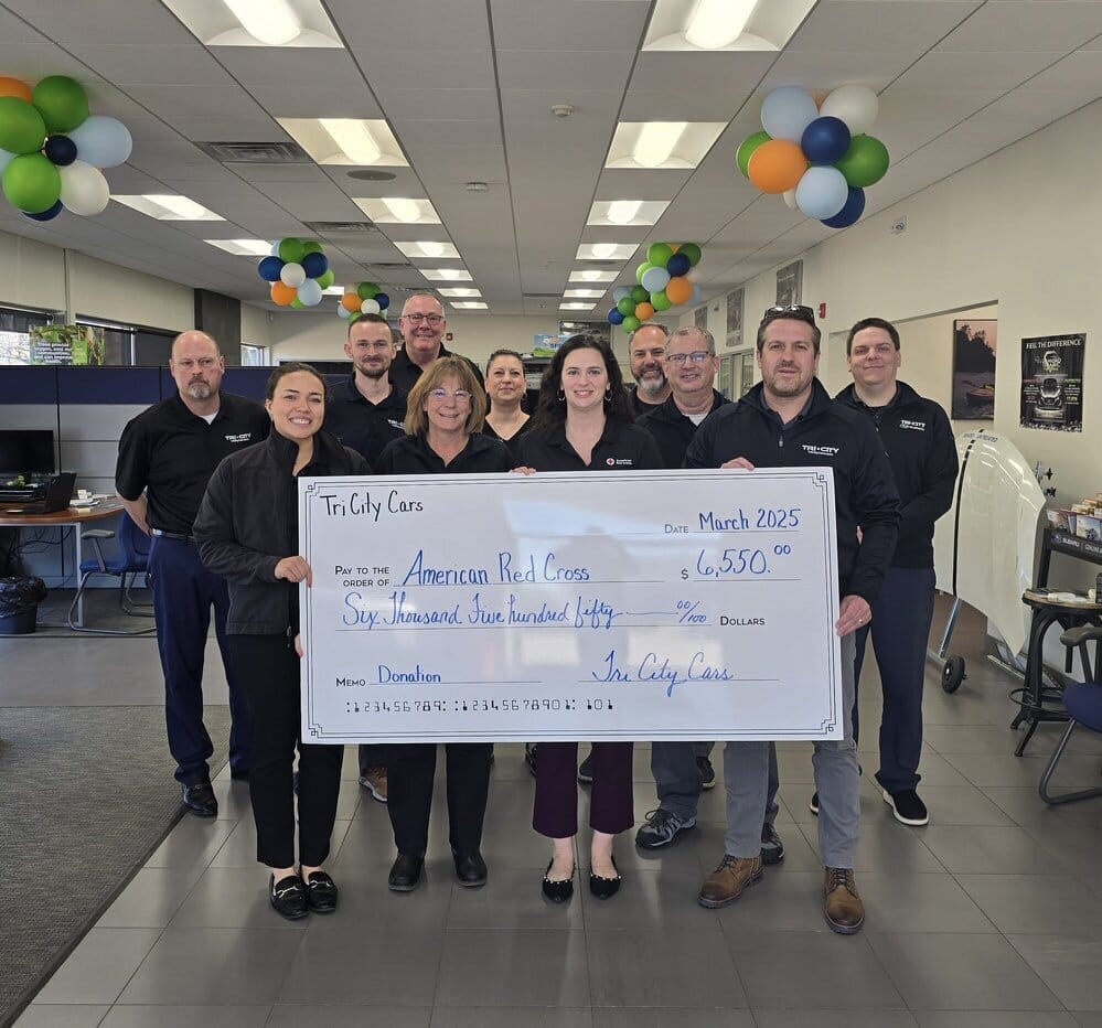 A group of people from Tri City Cars presenting a large donation check to the American Red Cross inside a dealership showroom decorated with balloons.