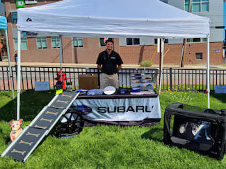 Man at an outdoor community event booth under a tent, with pet-related items like a ramp and crate displayed.