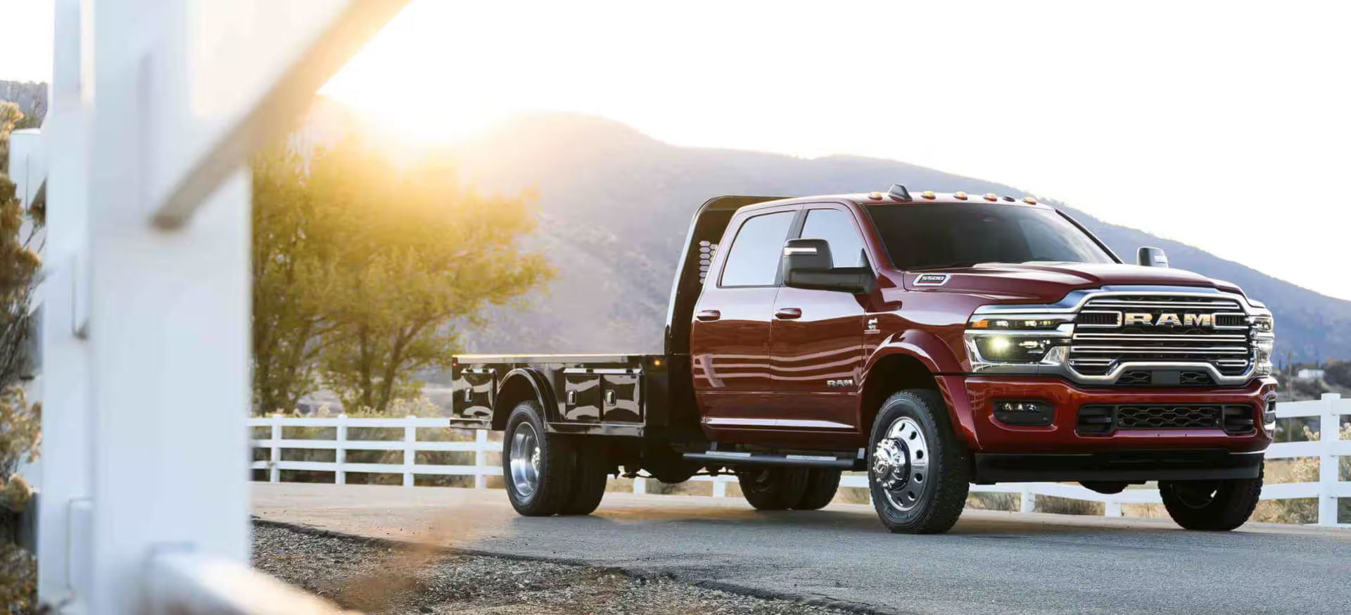 A red Ram heavy-duty pickup truck with a flatbed is parked on a rural road near a white wooden fence at sunset, with mountains and trees in the background.
