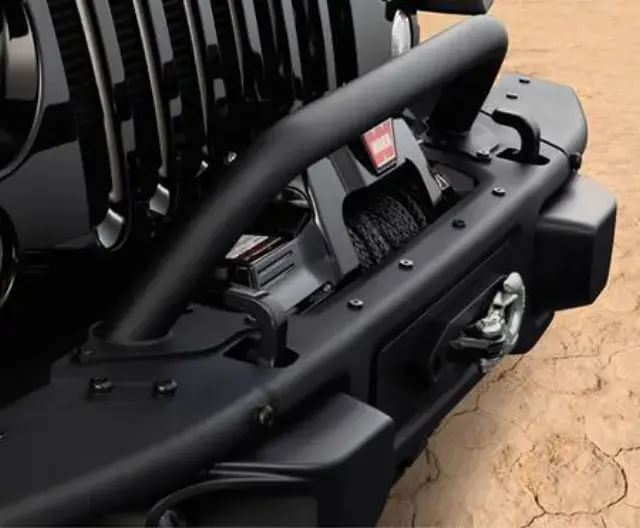 Close-up view of the front end of a black off-road vehicle, showcasing a heavy-duty bumper with a mounted WARN winch and tow hook, parked on cracked desert ground