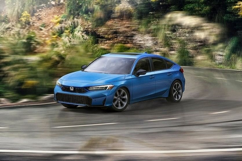 A blue 2025 Honda Civic Hatchback speeding around a winding road, surrounded by lush greenery and rocky terrain, showcasing dynamic motion and design.