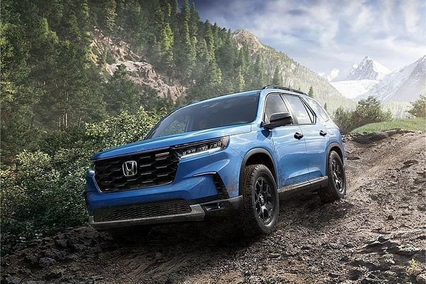 A blue 2025 Honda Pilot navigating a rugged, dirt trail surrounded by lush trees and mountains under a clear sky.