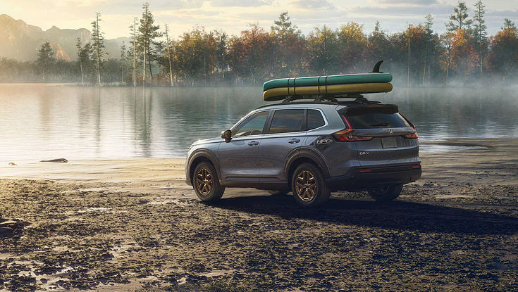 A gray Honda CR-V with a kayak and surfboard on its roof, parked on the shore of a lake.