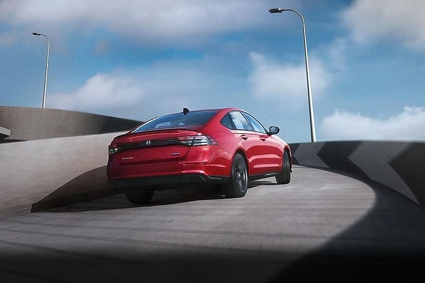 A sleek red 2025 Honda Accord Hybrid navigates a curved road under a cloudy sky, showcasing its sporty design and modern features.
