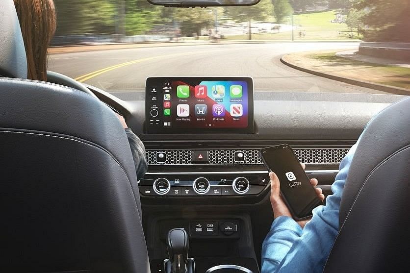 A view from the rear seat of a 2025 Honda Civic Hatchback, showcasing a digital dashboard and a passenger holding a smartphone with the CarPlay interface.