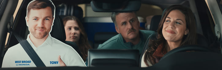 A family of three sits in a car, with a cardboard cutout of a man wearing a "West Broad Honda" shirt in the front passenger seat. The woman driving smiles, conveying a lighthearted mood.