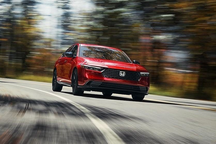 A sleek red 2025 Honda Accord Hybrid car maneuvers a curved road, set against a backdrop of colorful autumn foliage, showcasing speed and elegance.