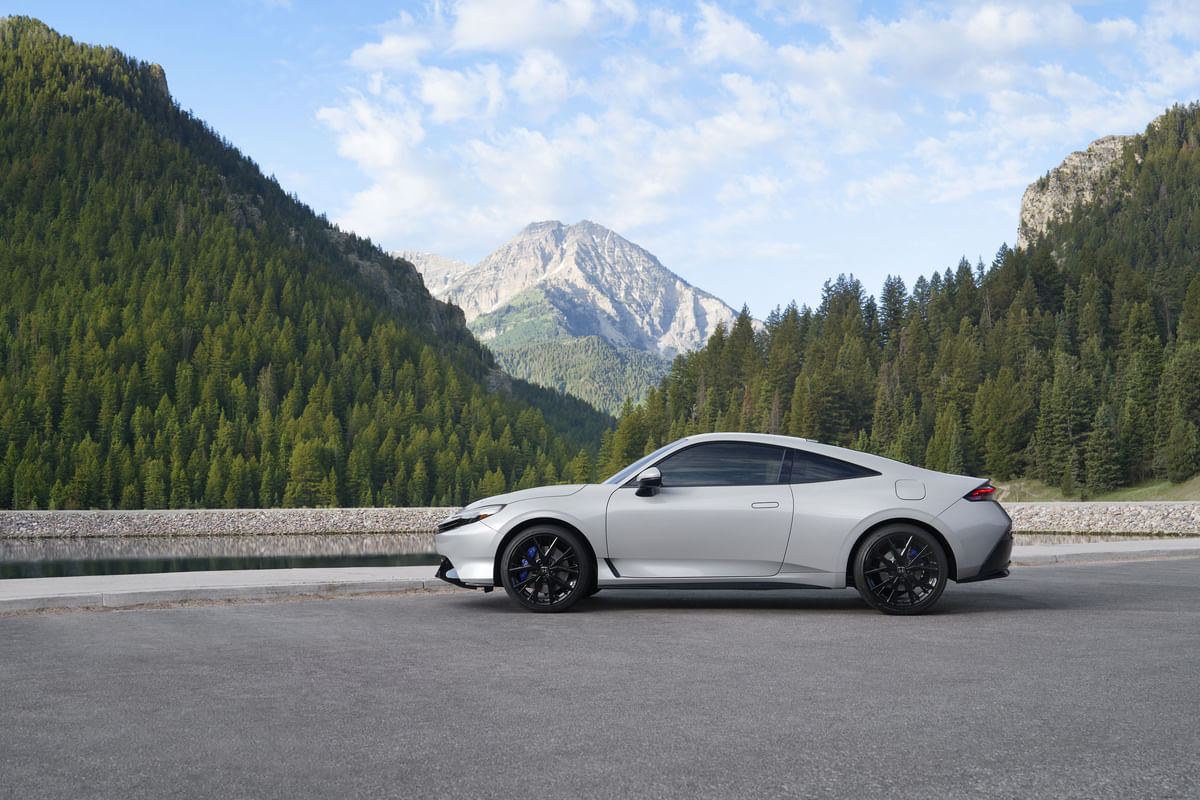 The side profile of a silver 2026 Honda Prelude parked on a road, showcasing its sleek coupe design and black alloy wheels against a backdrop of mountains and a forest-lined lake.