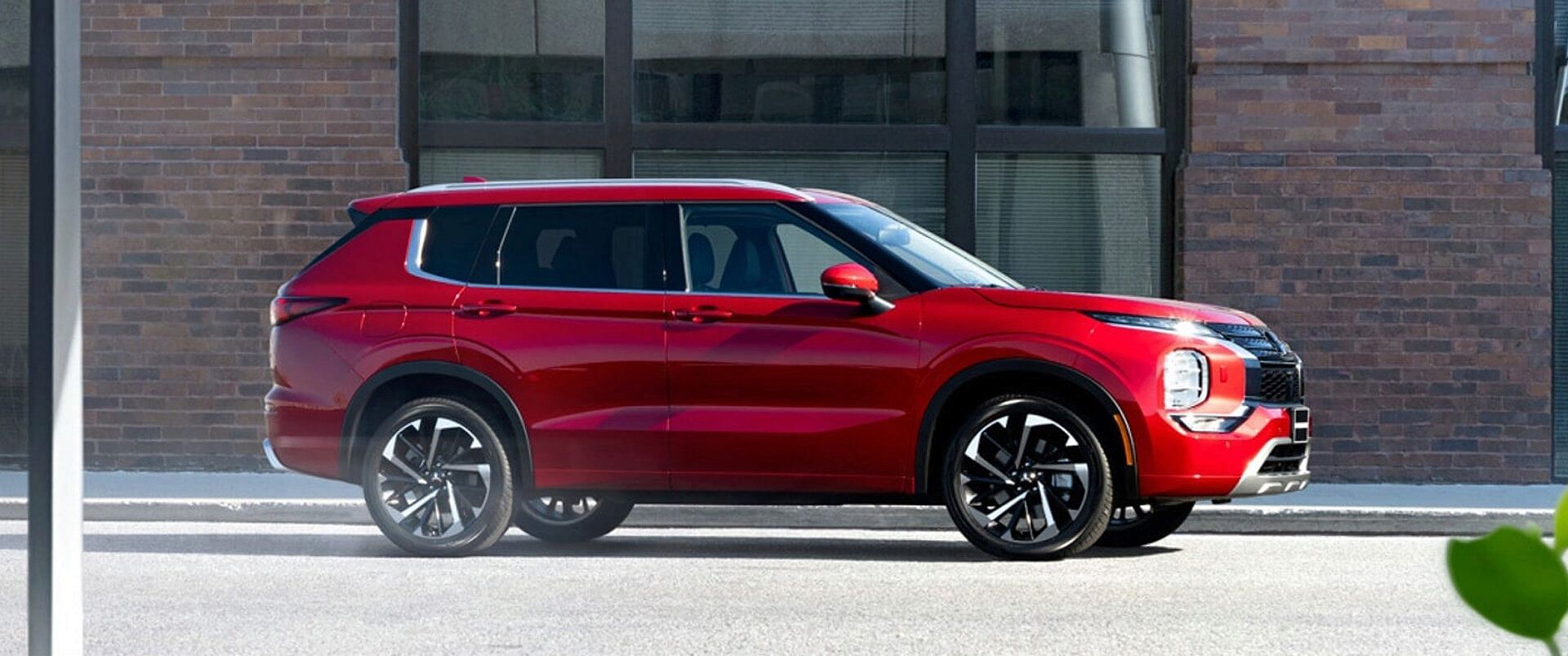 A red 2023 mitsubishi outlander standing on the street. Red brick building with a large window on the middle in the background