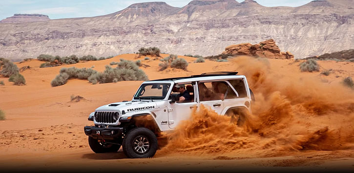 A white 24 Jeep Wrangler Rubicon driving off-road in the desert, kicking up sand as it goes.