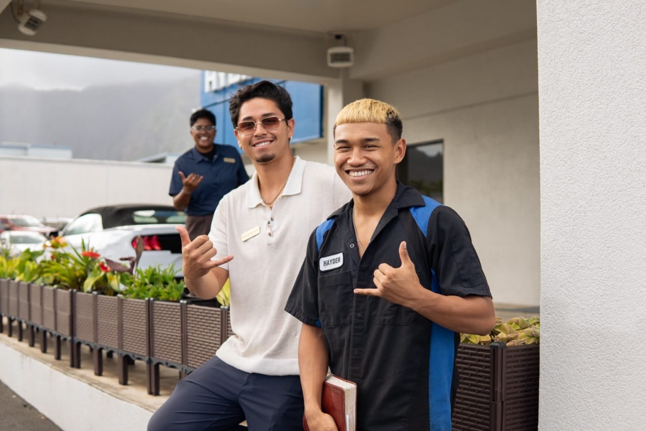 Two men stand side by side, smiling for a photo in front of a car dealership filled with vehicles.