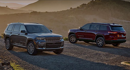 A gray 2024 Jeep Grand Cherokee Summit Reserve and a red Jeep Grand Cherokee Limited parked opposite of each other on a gravel mountain road at sunset.