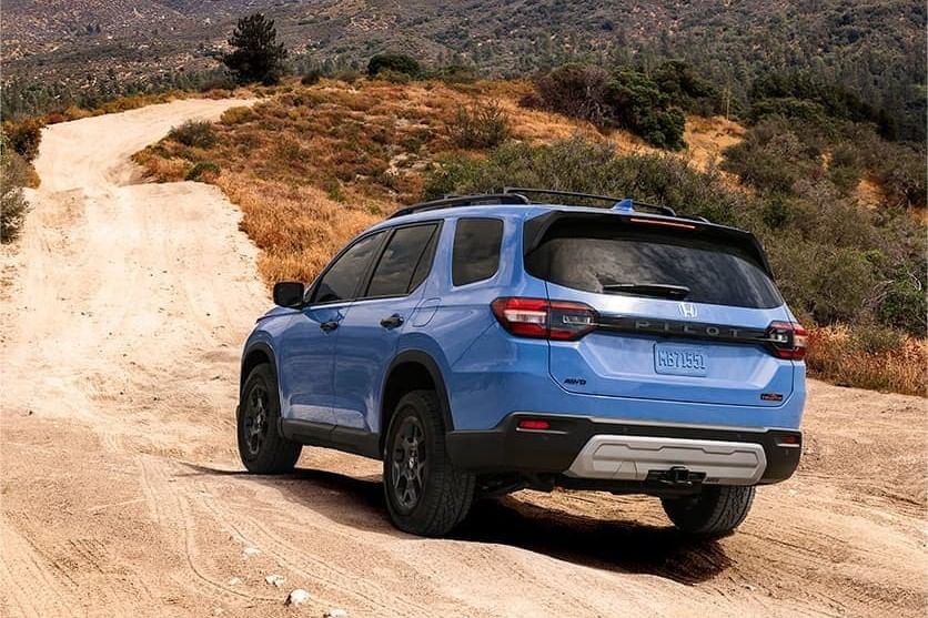 A blue 2025 Honda Pilot on a dirt road, surrounded by hills and greenery, ready for adventure in a rugged landscape.