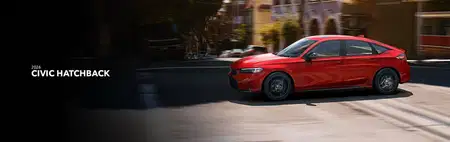 A red 2026 Honda Civic Hatchback speeds down a sunlit urban street, showcasing sleek design and dynamic motion. Bold, modern architecture blurs in the background.